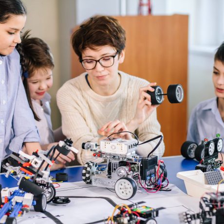 Curious clever pupils with the help of their female teacher doing a group project programming homemade robot using laptops on extracurricular classes